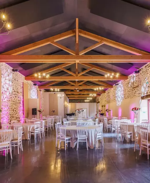 Salle de réception du Château de Gorze en configuration banquet – éclairage rose, mur en pierre apparente et charpente en bois