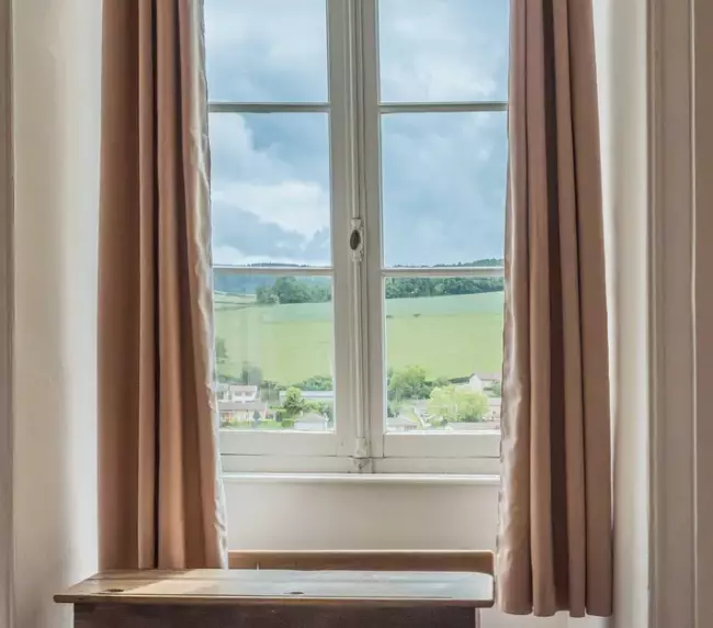 Vue sur la campagne depuis une fenêtre ancienne du Château de Gorze – pupitre en bois et rideaux beige